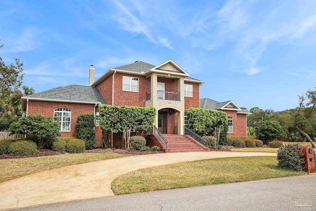 view of front of house featuring a balcony, brick siding, driveway, a chimney, and a front yard