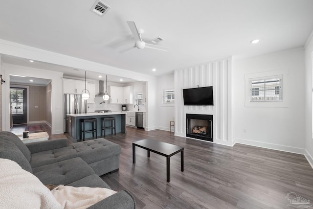 living room with sink, dark wood-type flooring, ceiling fan, ornamental molding, and a large fireplace