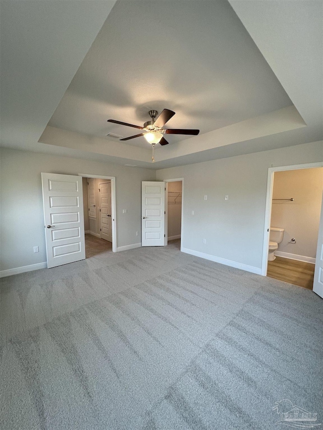 unfurnished bedroom with ceiling fan, ensuite bathroom, a spacious closet, light colored carpet, and a raised ceiling