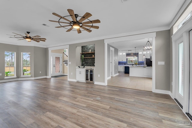 unfurnished living room featuring wine cooler, ceiling fan with notable chandelier, light hardwood / wood-style floors, and wet bar