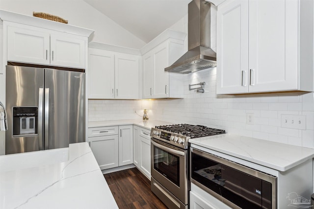 kitchen with lofted ceiling, wall chimney exhaust hood, light stone countertops, white cabinetry, and appliances with stainless steel finishes