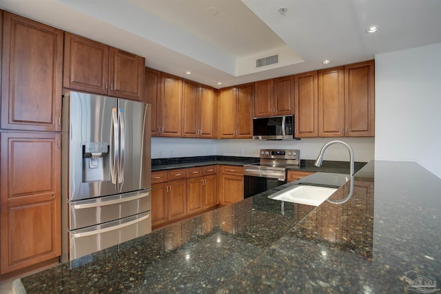 kitchen with appliances with stainless steel finishes, a tray ceiling, sink, and dark stone countertops