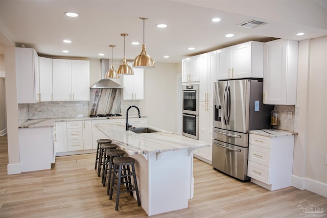 kitchen with wall chimney exhaust hood, white cabinetry, stainless steel appliances, sink, and a kitchen island with sink