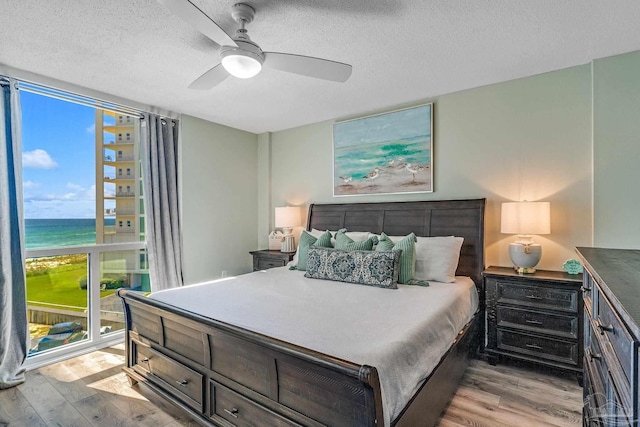 bedroom featuring a water view, light hardwood / wood-style flooring, a textured ceiling, and ceiling fan