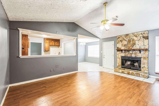 unfurnished living room with a textured ceiling, ceiling fan, light hardwood / wood-style flooring, a stone fireplace, and lofted ceiling