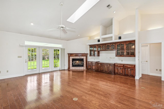 unfurnished living room with ceiling fan, a skylight, high vaulted ceiling, a tile fireplace, and hardwood / wood-style floors