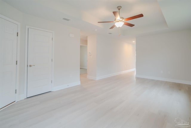 empty room with visible vents, a tray ceiling, light wood-style floors, baseboards, and ceiling fan