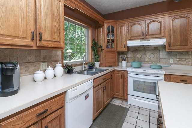 kitchen featuring light tile patterned floors, under cabinet range hood, white appliances, a sink, and brown cabinets