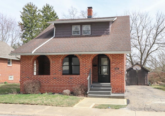 view of front facade featuring brick siding, a shingled roof, a shed, a chimney, and an outdoor structure