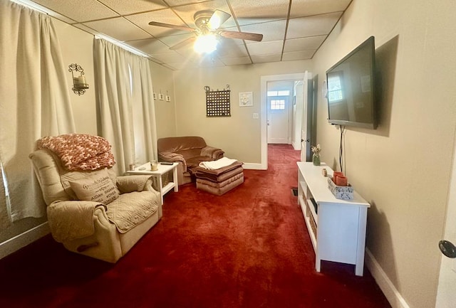 living area with a drop ceiling, dark carpet, and ceiling fan