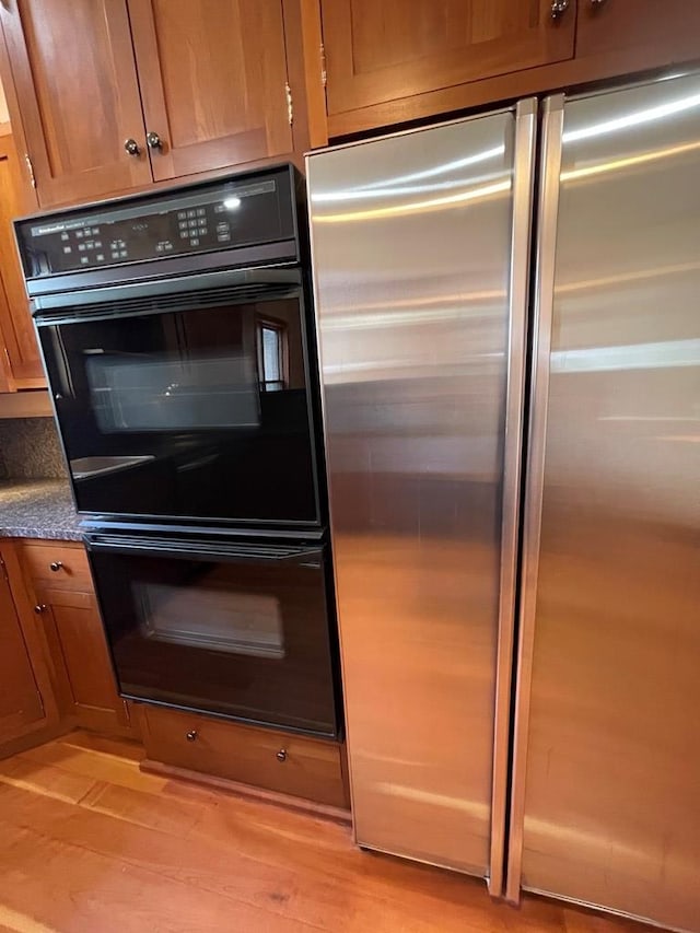 kitchen with light wood-style flooring, dobule oven black, dark countertops, stainless steel refrigerator, and brown cabinetry