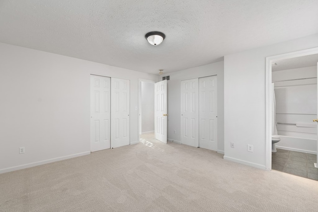 unfurnished bedroom featuring multiple closets, ensuite bathroom, light colored carpet, and a textured ceiling