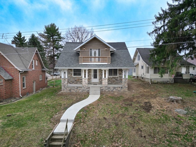 view of front of property with covered porch, stone siding, roof with shingles, a balcony, and a front lawn