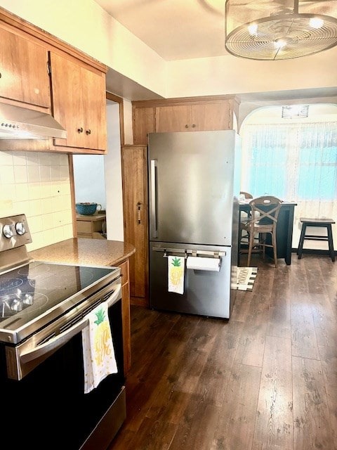 kitchen with brown cabinets, appliances with stainless steel finishes, arched walkways, decorative backsplash, and dark wood-style floors