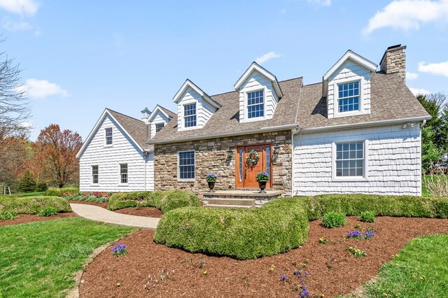 cape cod house with a chimney, roof with shingles, stone siding, and a front yard