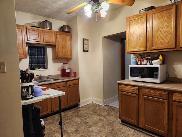 kitchen featuring a textured ceiling, white microwave, a sink, baseboards, and brown cabinetry