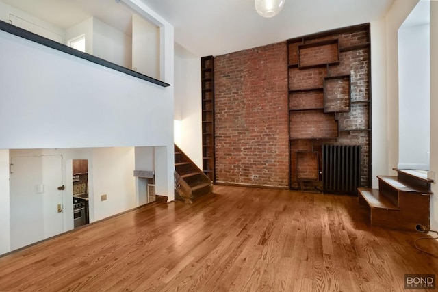 unfurnished living room with a brick fireplace, a high ceiling, brick wall, wood-type flooring, and radiator