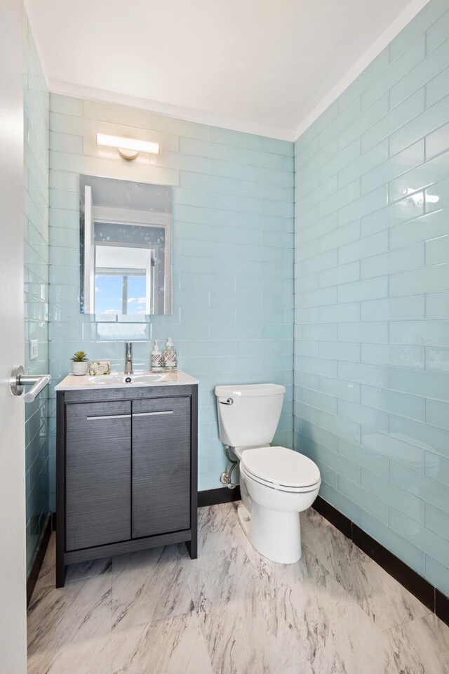 bathroom with toilet, marble finish floor, crown molding, vanity, and tile walls