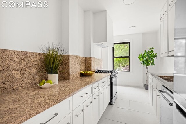 kitchen featuring wall chimney range hood, stainless steel range with gas stovetop, light tile patterned floors, white cabinets, and light stone counters