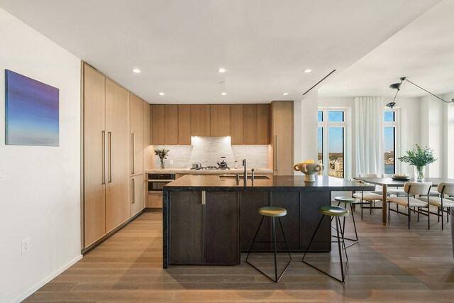 kitchen featuring hardwood / wood-style floors, backsplash, a kitchen island with sink, oven, and sink