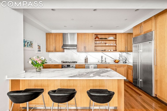 kitchen with wall chimney range hood, stainless steel appliances, sink, kitchen peninsula, and light wood-type flooring