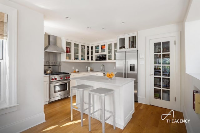 kitchen with premium appliances, a center island, light countertops, glass insert cabinets, and wall chimney range hood