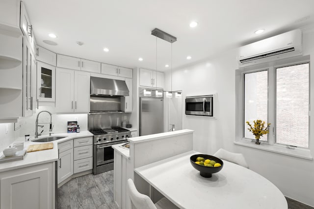 kitchen with an AC wall unit, wall chimney exhaust hood, white cabinetry, high end appliances, and sink