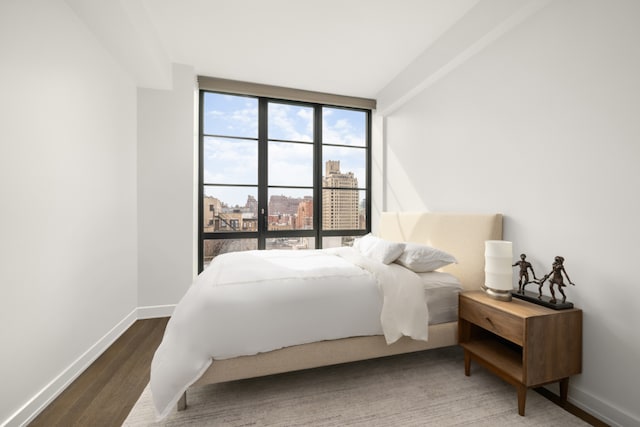 bedroom with dark wood-type flooring, a view of city, baseboards, and a wall of windows