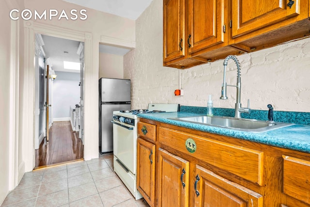 kitchen featuring a sink, freestanding refrigerator, brown cabinetry, light tile patterned floors, and white range with gas stovetop