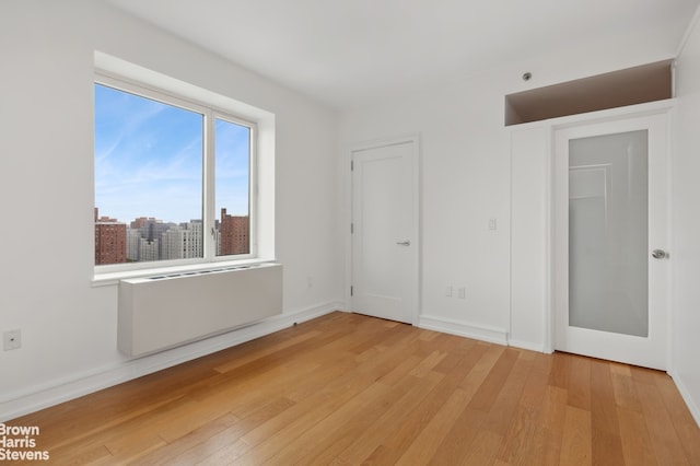 unfurnished bedroom with a city view, radiator, light wood-style floors, and baseboards