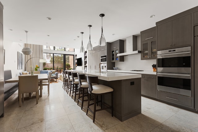 kitchen featuring a kitchen bar, decorative light fixtures, an island with sink, stainless steel double oven, and wall chimney range hood