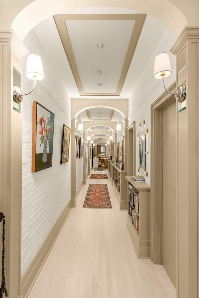 corridor with arched walkways, a tray ceiling, light wood-style floors, and ornate columns