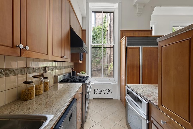 kitchen featuring appliances with stainless steel finishes, wall chimney exhaust hood, brown cabinetry, light tile patterned floors, and decorative backsplash