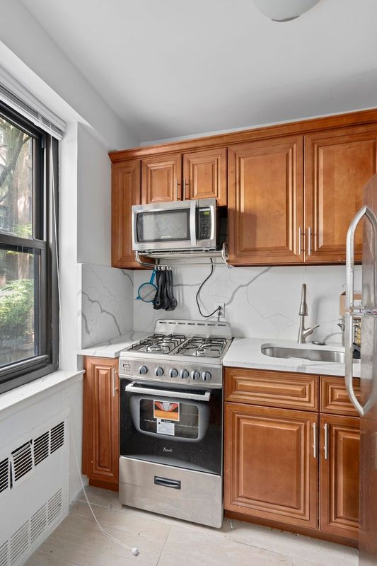 405 E 63rd St Unit 2B, New York City, NY 10065 - kitchen with stainless steel appliances, radiator, brown cabinetry, and light countertops