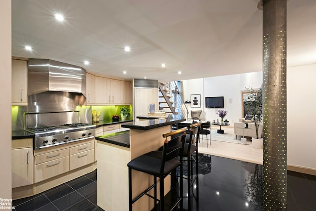 kitchen featuring a kitchen island, light brown cabinetry, wall chimney exhaust hood, a kitchen bar, and stainless steel gas stovetop