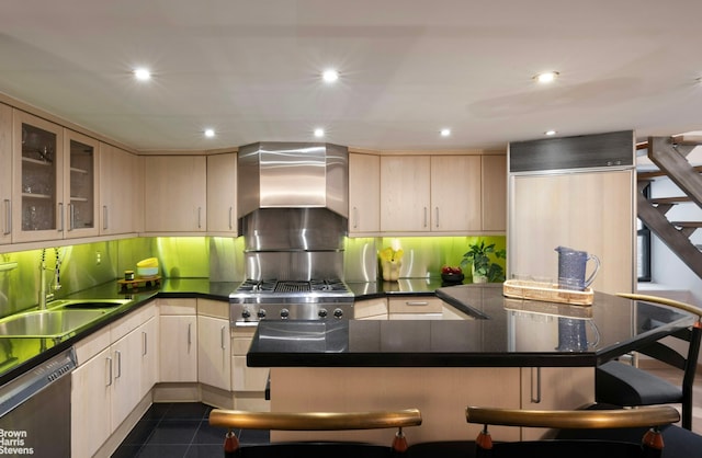 kitchen with light brown cabinets, a sink, a kitchen breakfast bar, stainless steel dishwasher, and wall chimney exhaust hood