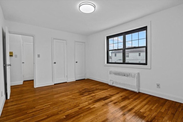 unfurnished bedroom with two closets, radiator heating unit, baseboards, and wood finished floors