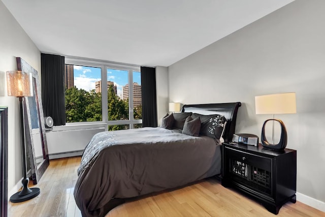 bedroom featuring light wood-style floors, a view of city, baseboards, and a baseboard heating unit