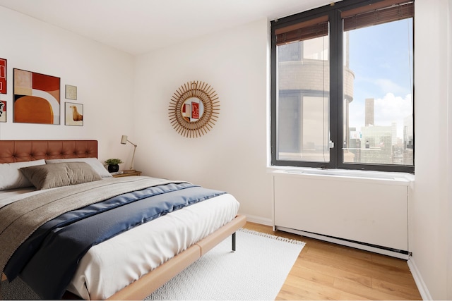 bedroom with a baseboard heating unit, a view of city, baseboards, and light wood-type flooring