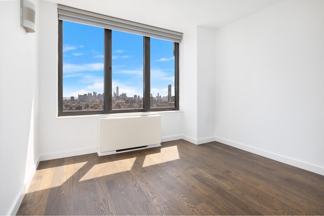 spare room featuring radiator heating unit, a view of city, baseboards, and dark wood-type flooring