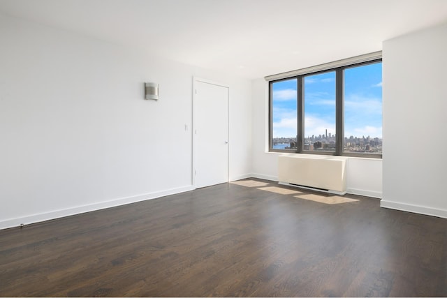 empty room with dark wood-style floors, baseboards, a city view, and radiator heating unit