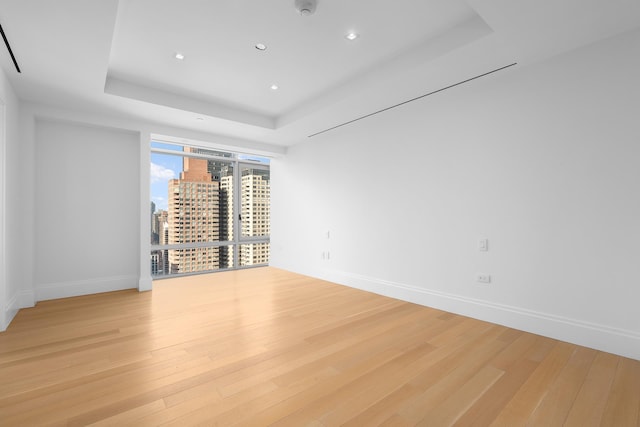 spare room with a tray ceiling, light wood-type flooring, a city view, baseboards, and recessed lighting