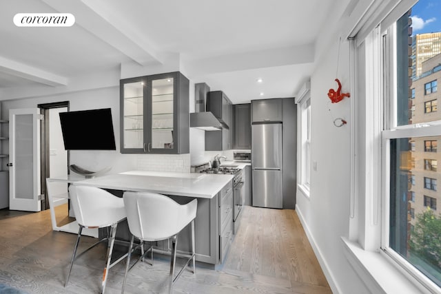 kitchen featuring appliances with stainless steel finishes, a breakfast bar, tasteful backsplash, gray cabinetry, and wall chimney range hood