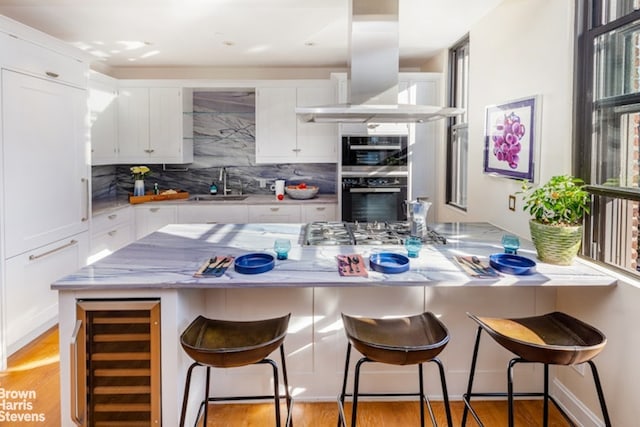 kitchen featuring double wall oven, island exhaust hood, wine cooler, white cabinets, and a breakfast bar