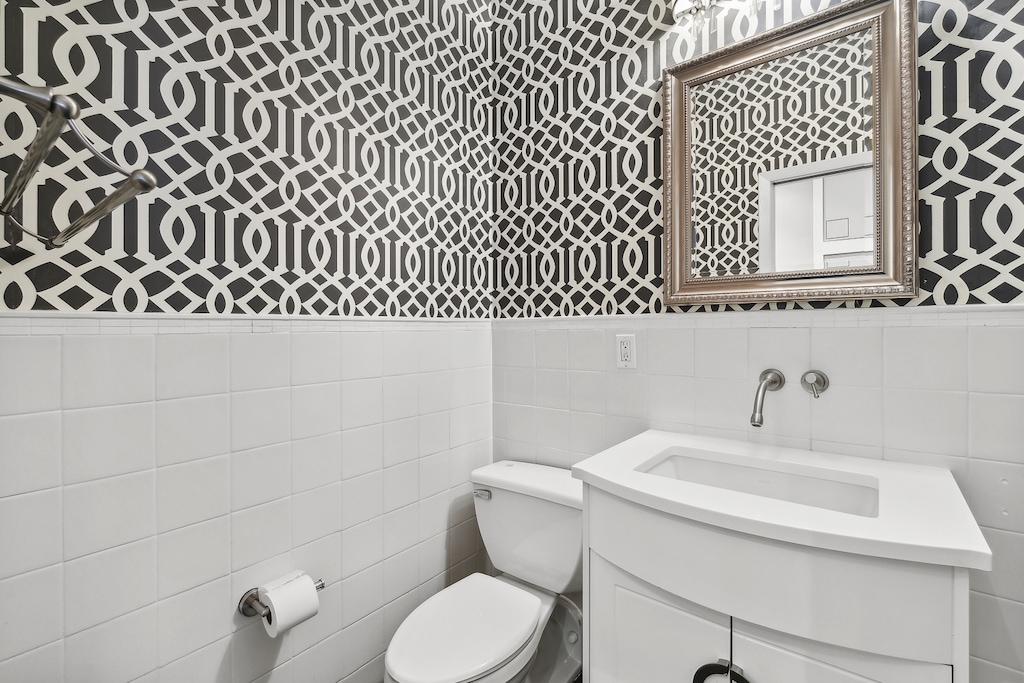 bathroom with wallpapered walls, toilet, tile walls, vanity, and a wainscoted wall