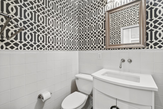 bathroom with wallpapered walls, toilet, tile walls, vanity, and a wainscoted wall