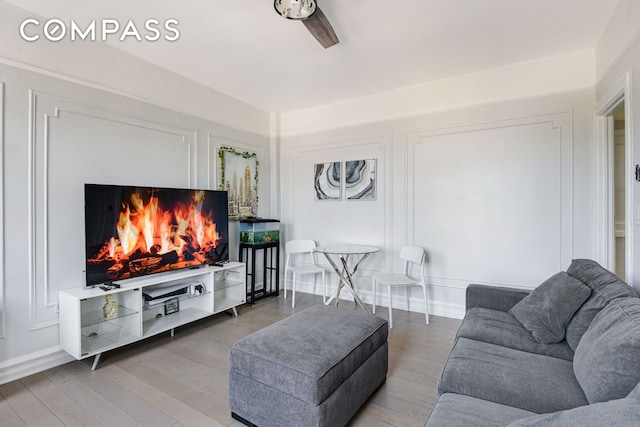 living room featuring a warm lit fireplace, ceiling fan, baseboards, and wood finished floors