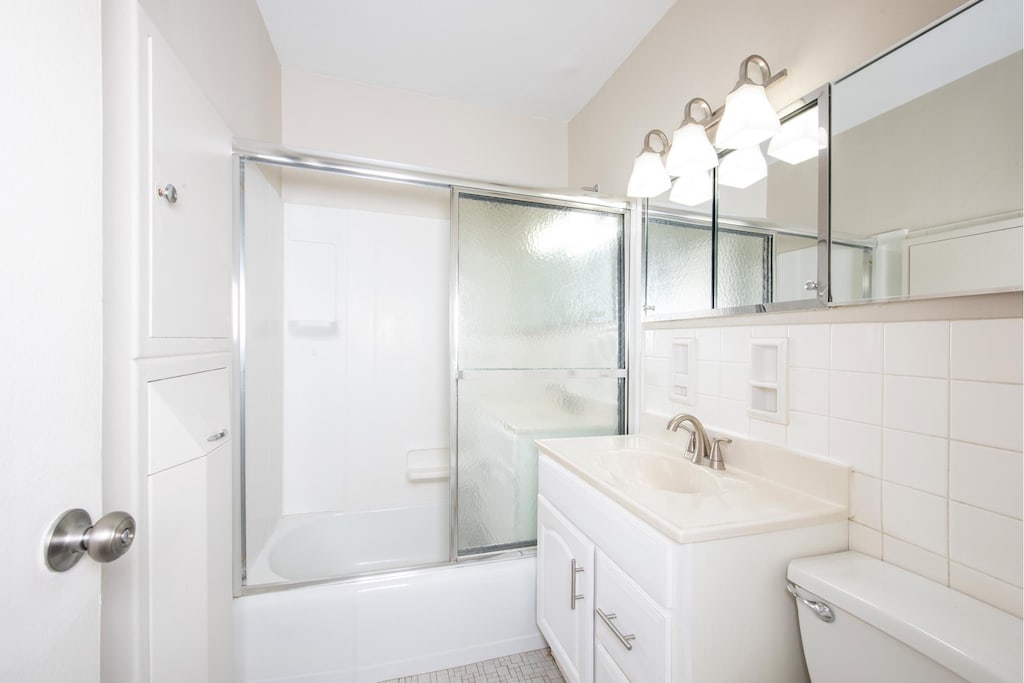 full bathroom featuring vanity, shower / bath combination with glass door, decorative backsplash, tile walls, and toilet