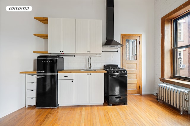 kitchen with radiator heating unit, white cabinetry, sink, black appliances, and wall chimney exhaust hood