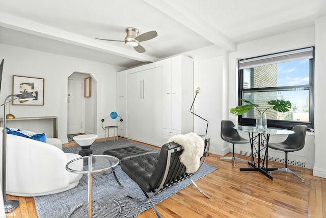 living room featuring baseboards, light wood-type flooring, beam ceiling, arched walkways, and a ceiling fan